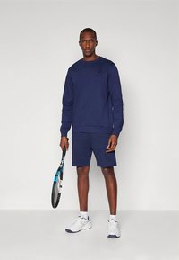 Tengerészkék pulóver és rövidnadrág szett puha anyagból, kerek nyakkivágással, bordázott mandzsettákkal és minimális logóval. Teniszütőt tart a kezében.