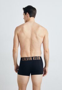 Schwarze Boxershorts mit breitem Bündchen, das "CALVIN KLEIN" in hellbraun zeigt. Glattes Material, enganliegendes Design und mittellange Beine.