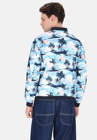 Jachetă bomber albastră cu un print tropical ce include palmieri și flori, cu guler și manșete negre contrastante, fabricată dintr-un material ușor.