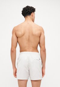 Pantaloni da bagno leggeri da uomo beige con vita elasticizzata e una tasca sul retro con un dettaglio del logo nero. Texture liscia.