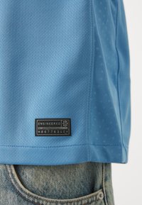 Maglietta sportiva azzurro chiaro realizzata in tessuto mesh traspirante, con un'etichetta nera in basso che riporta il testo "ENGINEERED" e un numero seriale.