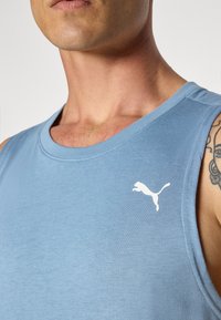 Lyseblå ærmeløs atletisk skjorte lavet af glat stof, med et hvidt Puma-logo på venstre brystområde. Nærbillede.
