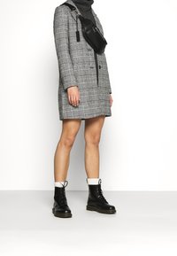 Persona vestida con un abrigo largo a rayas gris, un cuello de tortuga negro, una bolsa bandolera negra, calcetines blancos y botas negras con cordones de pie sobre un suelo blanco.