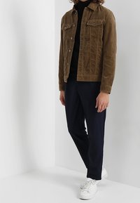 Veste en velours côtelé marron avec deux poches poitrine, portée sur un col roulé noir. Un pantalon bleu marine et des baskets blanches complètent la tenue.