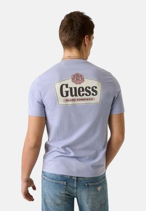 Uomo che indossa una maglietta Guess viola chiaro e jeans blu, di spalle, mostrando il logo e il nome del marchio sulla parte posteriore della maglietta.