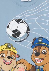 Lys blå T-skjorte med tegneseriehunder med gule og blå detaljer, en sort og hvit fotball, og hvite nettlinjer.