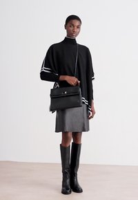 Poncho negru tricotat cu dungi albe, asortat cu o fustă din piele și cizme negre până la genunchi. Modelul poartă o geantă de mână structurată, neagră.