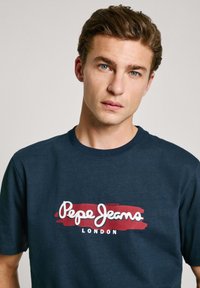 Jūras zila kokvilnas t-krekls ar baltu "Pepe Jeans" logotipu uz sarkanas otas triepuma fona. Īsas piedurknes un apaļa kakla izgriezuma forma.