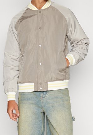 Homme portant une veste bomber beige et gris clair avec des poignets et un bas rayés blanc et jaune, associée à un jean bleu clair.
