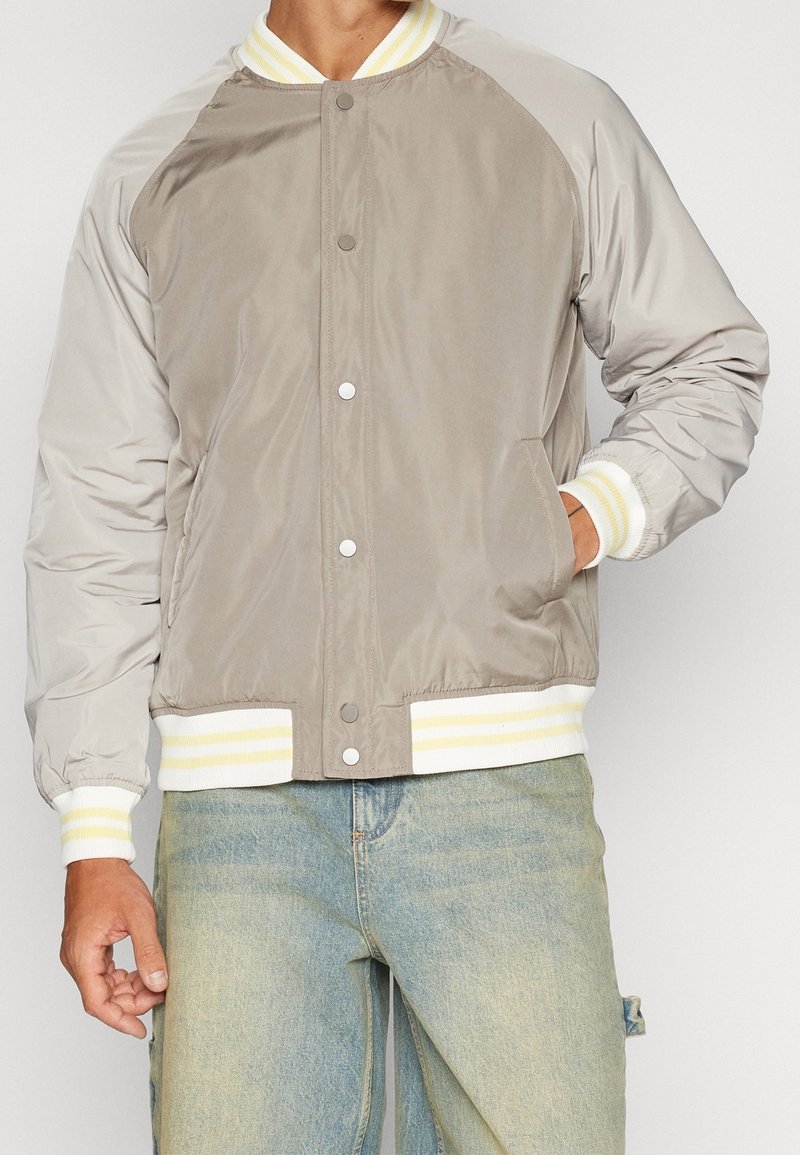 Homme portant une veste bomber beige et gris clair avec des poignets et un bas rayés blanc et jaune, associée à un jean bleu clair.