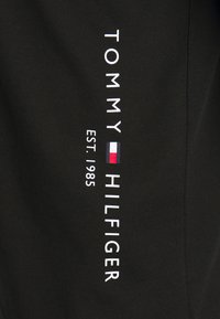 Black cotton fabric featuring a vertical white logo with "TOMMY HILFIGER" and "EST. 1985," alongside a red, white, and blue rectangular emblem.