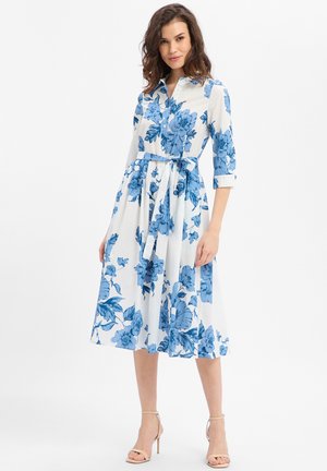 Vrouw draagt een witte midi-jurk met blauwe bloemenprint, driekwart mouwen, een kraag en een taillestrik, gecombineerd met naakte sandalen met hoge hakken.