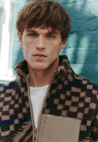 Fleece zip-up jacket featuring a brown and beige checker pattern, a tan panel, and a high collar. Worn over a white sweatshirt.