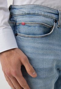 Světle modré džíny z denimu s jemnou texturou. Na zadním kapsu je červená etiketa a jeansy mají tradiční pětikapsový design.