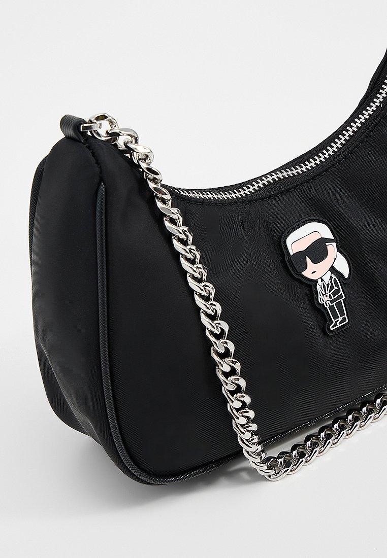 Sac à main en nylon noir avec une bandoulière en chaîne argentée, doté d'une fermeture éclair et d'un détail d'épingle de personnage de dessin animé. Texture lisse avec une forme arrondie.