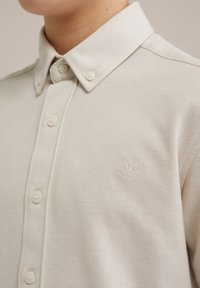 Camisa de color beige claro con cuello, confeccionada en tejido texturizado, que presenta una tapeta de botones y un sutil logo bordado en el pecho.