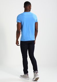 Helles blaues Baumwoll-T-Shirt mit rundem Ausschnitt und kurzen Ärmeln, kombiniert mit dunklen Jeans und grauen Sneakern, von hinten gesehen.