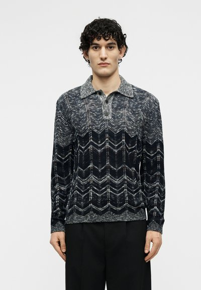 Νέος άνδρας με σγουρά μαλλιά, που φοράει ένα μακρυμάνικο σκούρο πουλόβερ πλεκτό πόλο με σχέδια και μαύρο παντελόνι, στέκεται μπροστά σε ένα απλό φόντο.