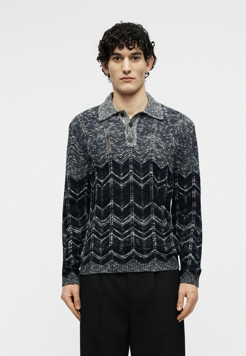 Young man with curly hair wearing a long-sleeve dark patterned knit polo shirt and black pants standing against a plain background.