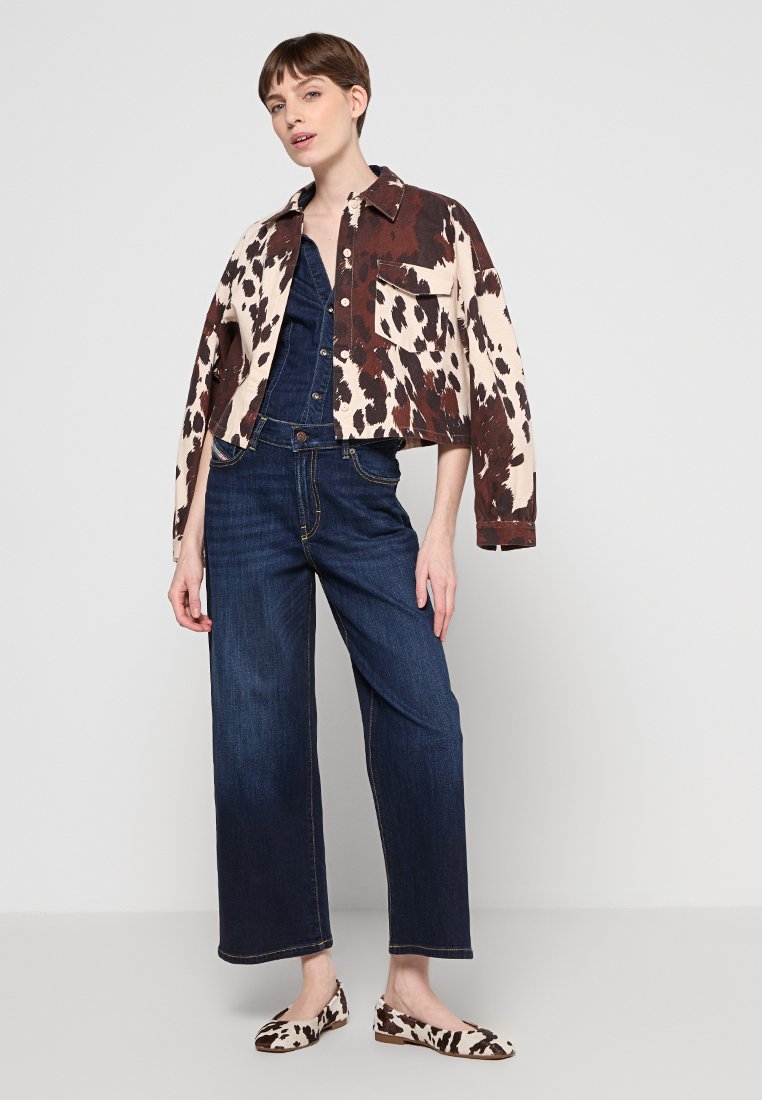 Cropped jacket with a brown and cream cow print, worn over a denim jumpsuit. Model also wearing dark blue wide-leg jeans and matching flats.