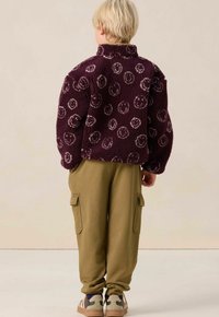 Bordeaux fleece jack met een smiley-gezicht patroon, hoge kraag en oversized mouwen, gecombineerd met olijfgroene cargo-broeken en gestreepte sneakers.