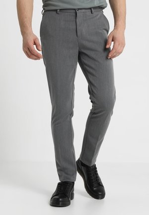 Uomo che indossa pantaloni grigi slim fit e sneakers nere, in piedi su uno sfondo chiaro e uniforme.