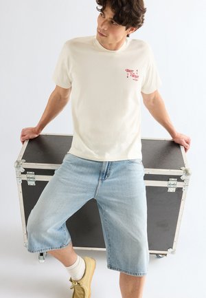 Mladý muž v bielom tričku s potlačou, svetlomodrých džínsových kraťasoch a béžových topánkach, opierajúci sa o čierny transportný kufor.