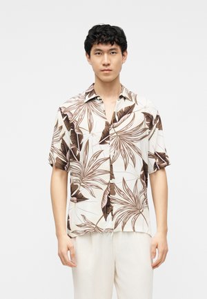 Άνδρας που φοράει λευκό κοντομάνικο πουκάμισο με μεγάλο καφέ τροπικό σχέδιο φύλλων και λευκό παντελόνι, στέκεται μπροστά σε απλό φωτεινό φόντο.