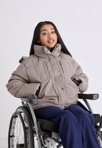 Puffer jacket in light taupe with a high collar, large buttons, and side pockets, worn over navy blue trousers, seated in a wheelchair.