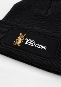 Black knit beanie featuring a patch with a cartoon character and the text "KLEINES SCHLITZOHR" in white. Textured ribbed fabric.
