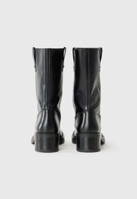 Botas de tobillo de cuero negro con un tacón grueso, que cuentan con una cremallera y pequeños lazos a los lados. Colocadas sobre un fondo neutro.