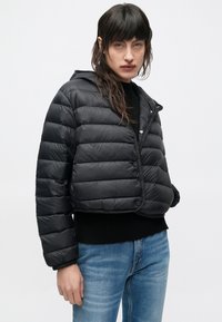 Γυναίκα που φοράει μαύρο κοντό μπουφάν με κουκούλα, μαύρο πουλόβερ και μπλε τζιν, στέκεται μπροστά σε απλό λευκό φόντο.