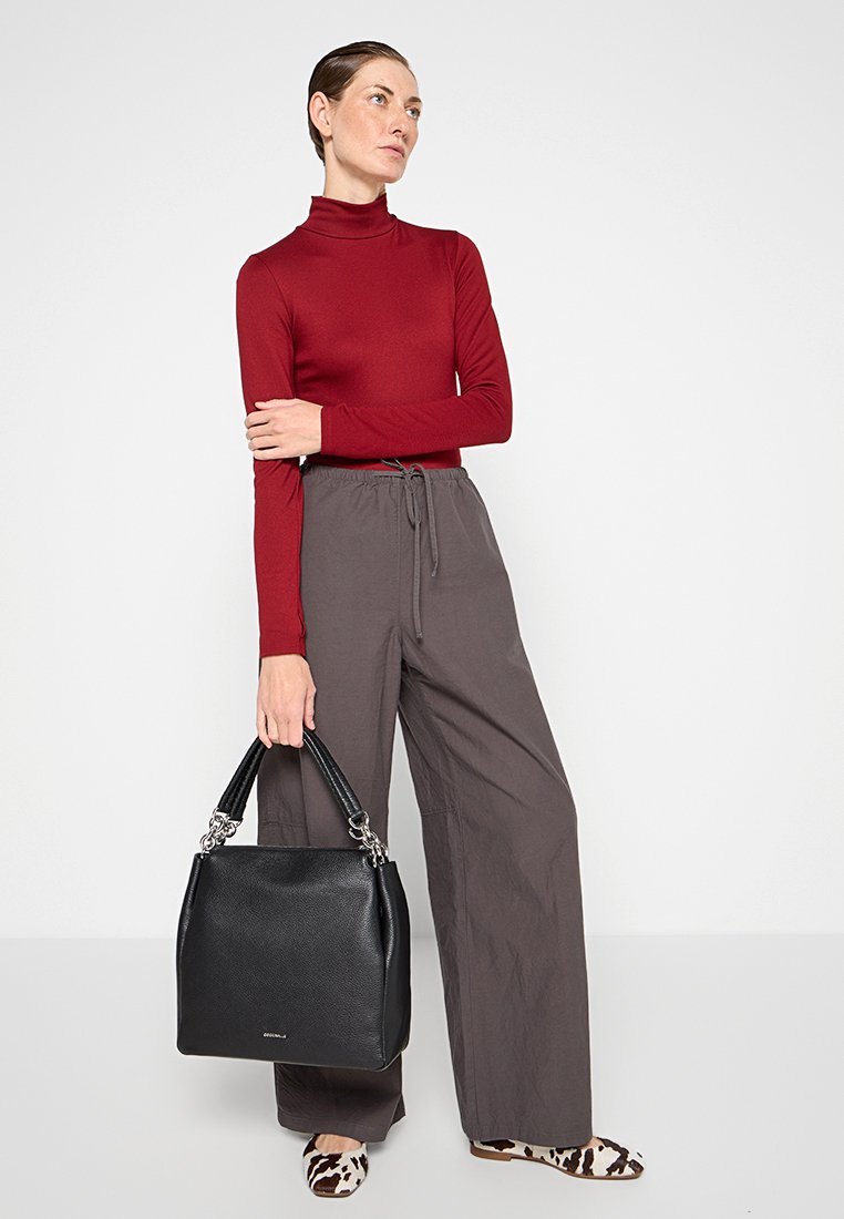 Borsa a mano nera in pelle con una forma strutturata, caratterizzata da una superficie testurizzata e hardware in argento. Donna in un dolcevita rosso e pantaloni larghi grigi.