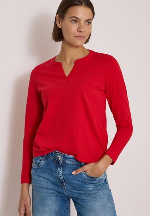 Femme portant un haut rouge à manches longues avec un petit col en V et un jean bleu, posant avec une main dans la poche devant un fond uni.