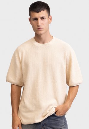 Νεαρός άνδρας με κοντά σκούρα μαλλιά, φοράει εκρού πλεκτό πουλόβερ με κοντά μανίκια, στέκεται με το ένα χέρι στην τσέπη μπροστά από φωτεινό φόντο.