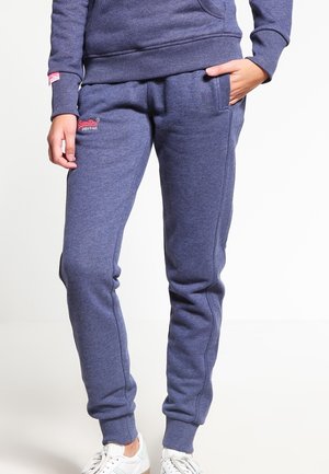 Person trägt blaue, schmal zulaufende Jogginghosen mit elastischen Bündchen und weißen Sneakers, Hände in den Taschen, vor einfachem Hintergrund.