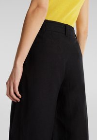 Femme portant un pantalon noir taille haute et un haut jaune sans manches, vue de dos avec la main reposant près de la hanche.