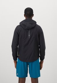 Sort vandtæt jakke med hætte, med et reflekterende logodesign og justerbar kant, parret med blå shorts. Glat, letvægtsstof.