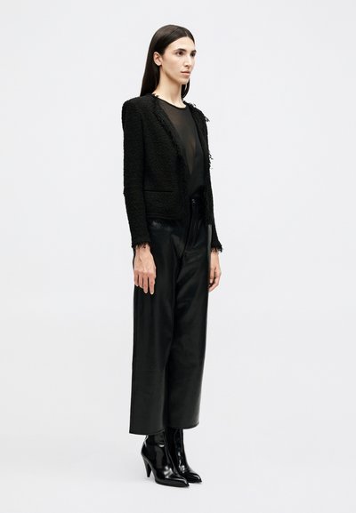 Žena stojaca, oblečená v čiernom textúrovanom blejzri, priesvitnom čiernom topu, voľných čiernych nohaviciach a čiernych členkových čižmách s vysokým podpätkom a špicatou špičkou.