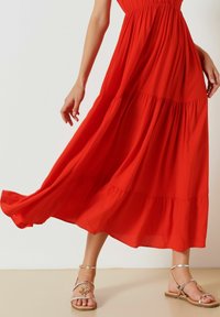 Robe maxi rouge en tissu léger avec des couches superposées, associée à des sandales métalliques avec des accents décoratifs et des sangles fines.