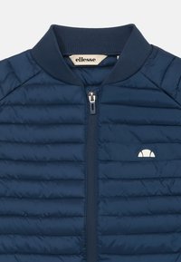 Marinblå vadderad jacka med ribbat krage, framzipper och liten vit logotyp på vänster bröst. Har horisontella sömmar.