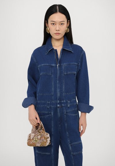 Denim overal s uzáverom, s rolovanými rukávmi a detailmi vrecka. Skombinovaný s malou, svetlou kabelkou s vzorom.