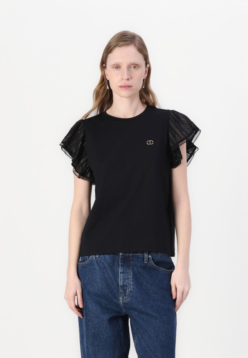 Zwart T-shirt met korte, gerimpelde doorschijnende mouwen met metallic draden. Gouden logo-accent aan de voorkant. Draagt in combinatie met high-waisted jeans.
