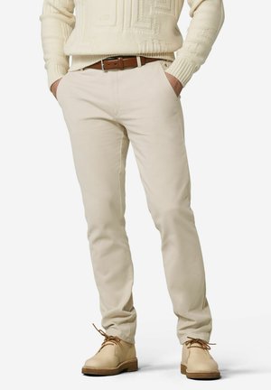 Mann trägt beige Slim-Fit-Hosen, hellbraunen Ledergürtel, cremefarbenen Strickpullover und beige Schnürschuhe mit braunen Sohlen. Hände in den Taschen.