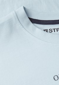 Světle modré bavlněné tričko s kulatým výstřihem, hladkou texturou a jemným logem. Výstřih má kontrastní tmavý lem.