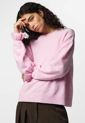 Jeune femme aux cheveux bouclés portant un pull rose avec des poignets volantés, posant la main sur le front devant un fond uni.