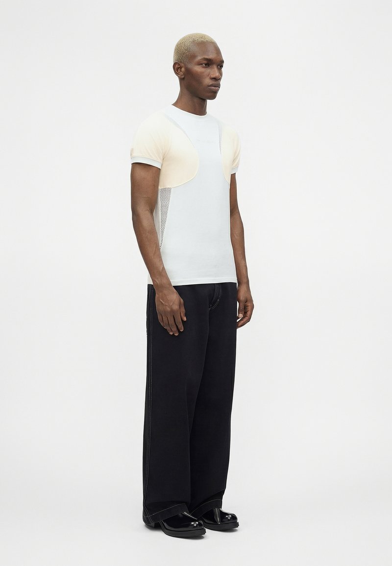 Homme aux cheveux blonds courts portant un T-shirt blanc moulant avec des manches courtes beige clair, un pantalon noir à jambes larges et des chaussures noires.