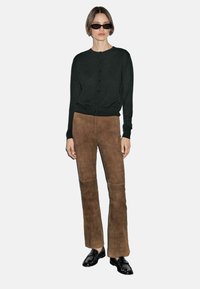 Model in black cardigan and brown suede pants poses against a plain background, wearing sunglasses and black shoes, with hands relaxed at her sides.