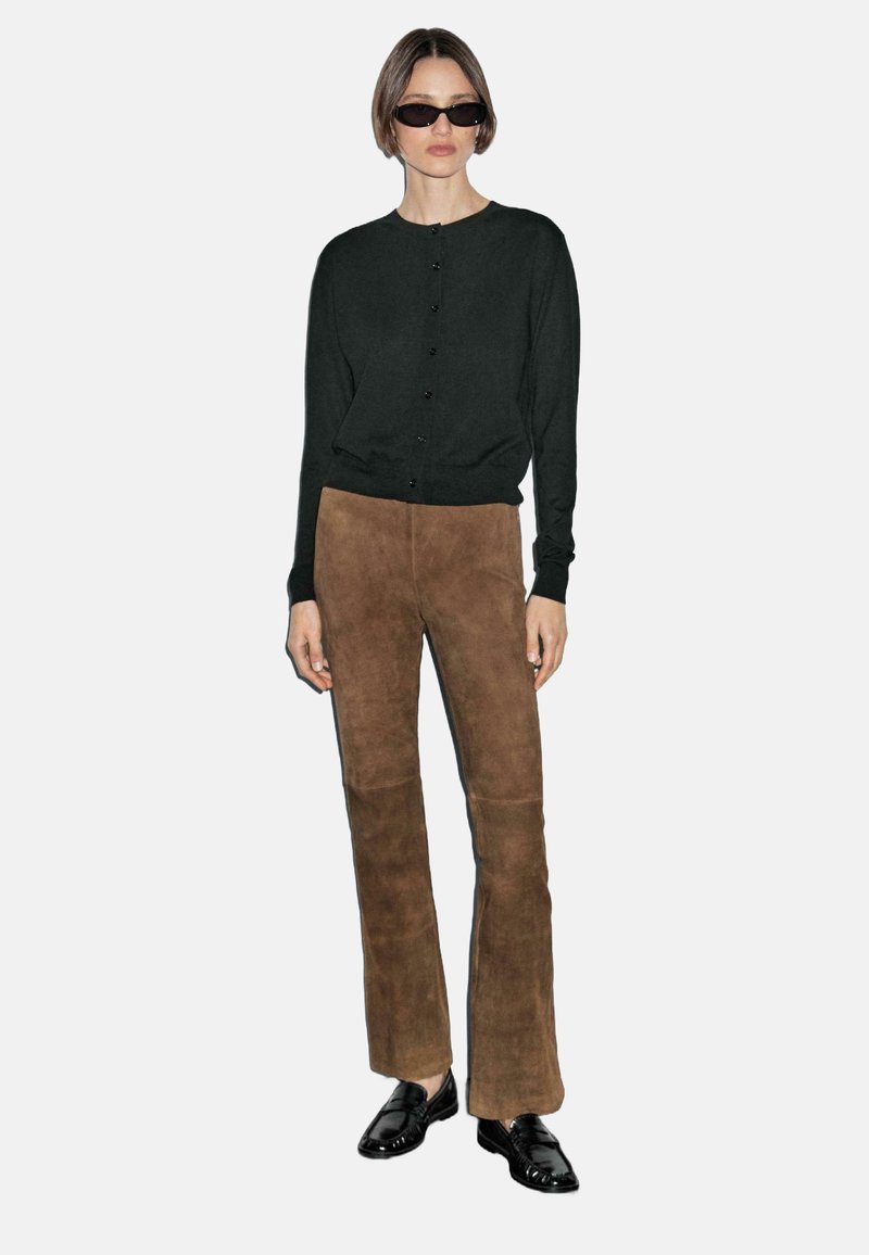 Model in black cardigan and brown suede pants poses against a plain background, wearing sunglasses and black shoes, with hands relaxed at her sides.