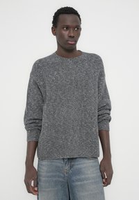 Άνδρας που φοράει γκρι πουλόβερ με ριμπ ύφανση και μανίκια γυρισμένα, και ξεθωριασμένο μπλε τζιν, στέκεται μπροστά σε απλό λευκό φόντο.